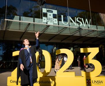 A man in graduation regalia standing in front of a sign that says 2023 throw his graduation cap in the air