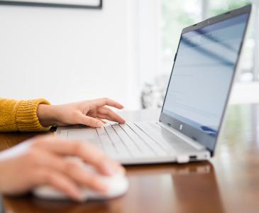 Close up of a laptop on a wooden table and a woman in a mustard sweater with her hands on the mouse