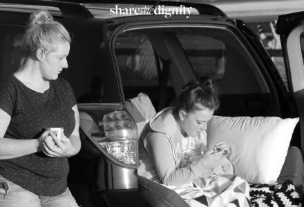 A woman stands beside the open boot of a car where a girl sits with a pillow and blanket
