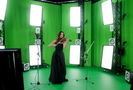 A woman in a long black dress playing violin in a green screen studio with multiple cameras and lights