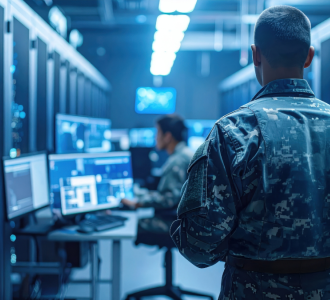 A man in an army uniform walking into a data centre with another man working on a computer.