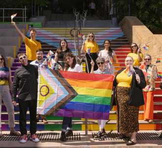 A group of people gathered on the Basser steps with a Progress Pride flag