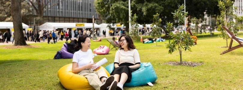 Two women sit laughing together on colourful beanbags on the lawn in front of a building