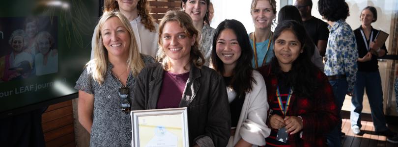 A group of seven scientists smile in front of a building, holding a framed award
