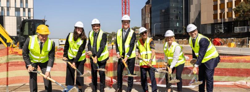 Seven men and women wearing hard hats and hi-res vests shovel earth from the ground of a construction site