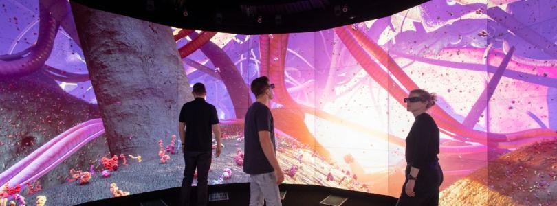 A blonde woman wearing a virtual reality headset and two men stand in front of a colourful circular life-size screen